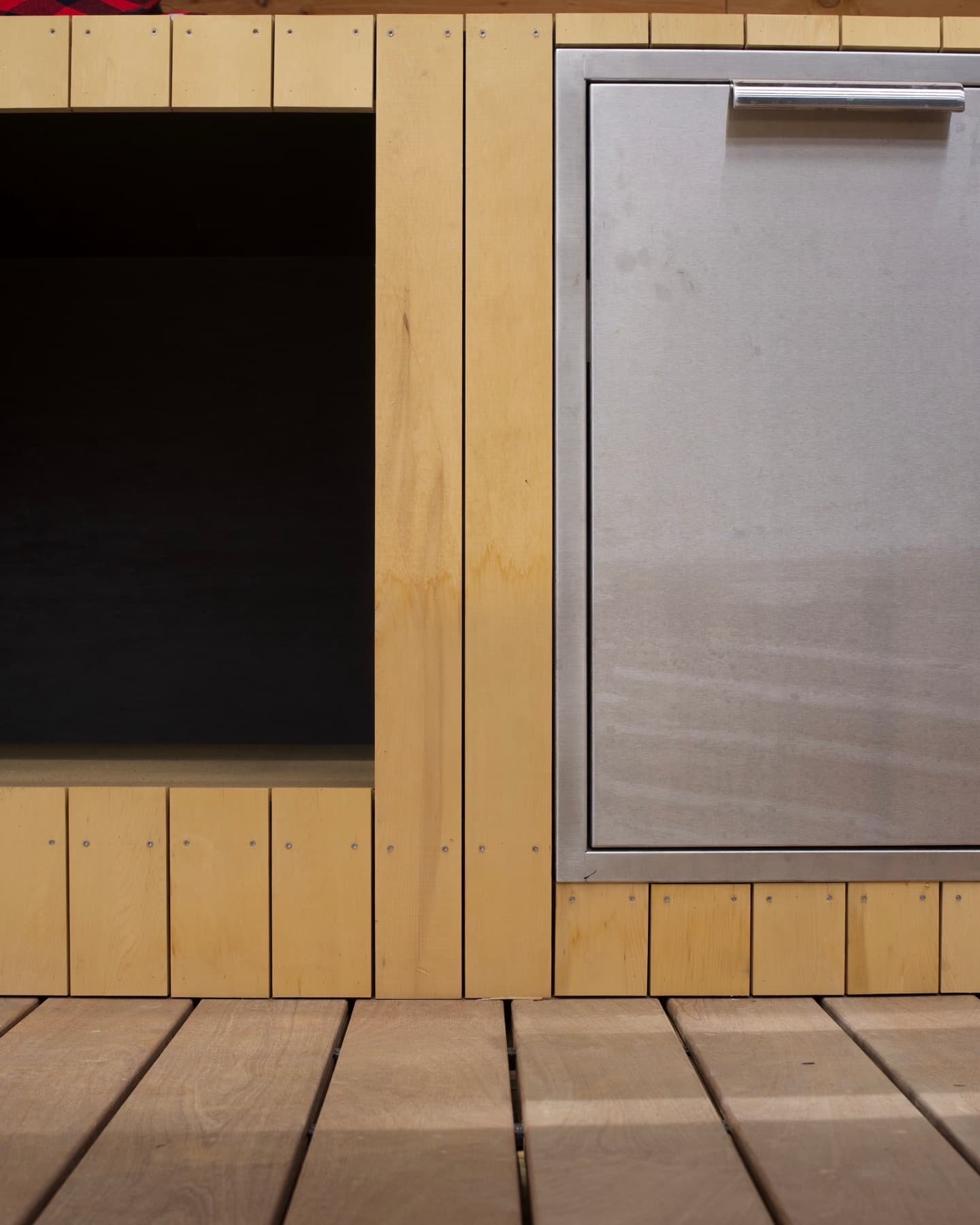 Outdoor kitchen detail: pale wood plank cladding around a stainless steel access door over ipe deck boards