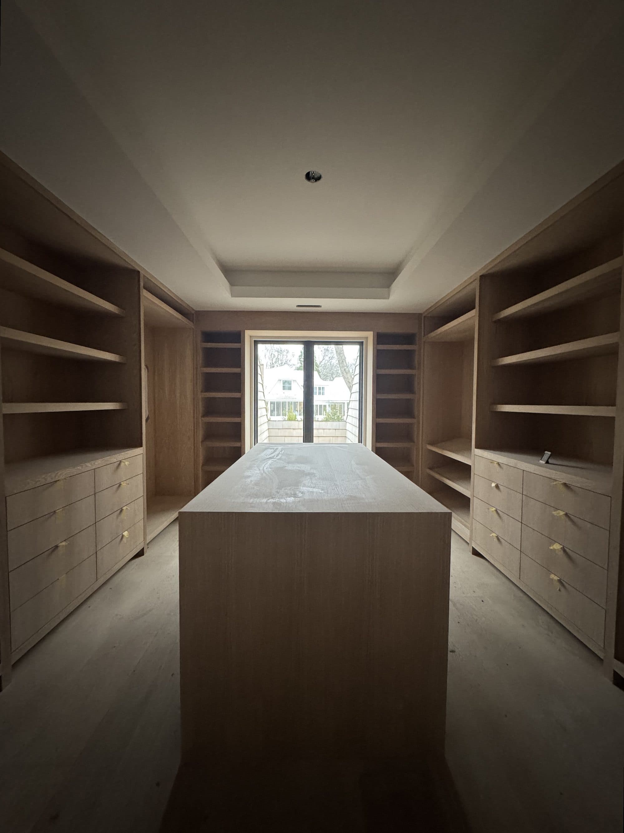 Custom white oak dressing room built by Master Team Construction: fitted shelving and drawers on both walls around a wood-wrapped island, lit by a single window