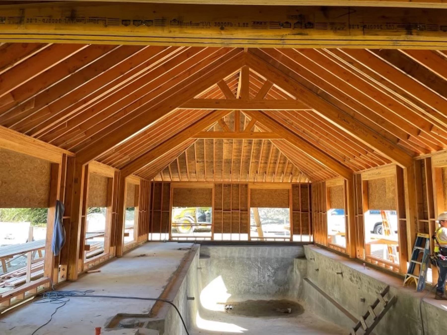Interior of a pool house under construction with a cathedral of exposed rafters above an empty gunite pool