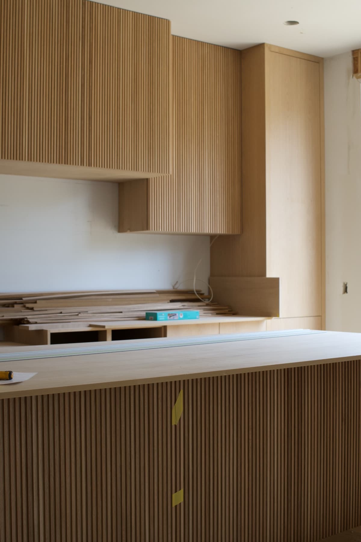 White oak kitchen under construction with fluted cabinet fronts on the upper cabinets and island, stacks of oak boards on the counter