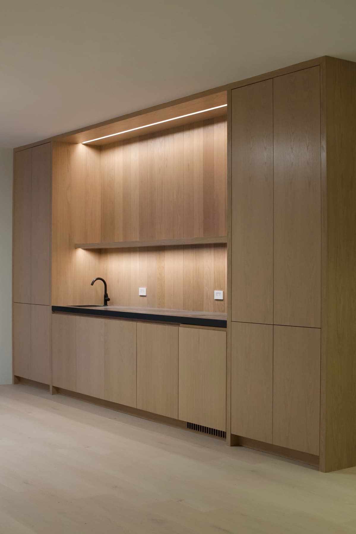 Full-height white oak wet bar wall with black stone counter, black faucet, and warm under-shelf lighting